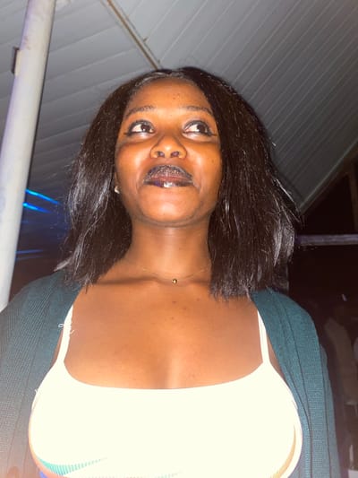 A medium shot shows a young Black woman looking upwards and to her right, with her mouth slightly open and her eyes wide. She has dark, shoulder-length hair, dark eyeliner, and dark lipstick with a glittery sheen. She is wearing a dark green cardigan over a white tank top with thin, multicolored stripes on the strap. A thin gold necklace with a small gold ball pendant is visible around her neck. The background consists of a white ceiling with parallel grooves, a white pole on the left, and a dark, blurred background with hints of blue lights on the left side, suggesting an indoor or covered outdoor setting. The overall lighting is dim, with the woman's face illuminated by a light source from above and slightly to her left. The image appears to be taken at night, possibly in a social gathering or entertainment venue in Wumat, Nigeria.