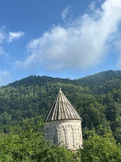 The image shows a stone bell tower of the Teghut Monastery in Teghut, Armenia, set against a backdrop of a lush, green, tree-covered mountain under a blue sky with white clouds. The bell tower has a conical roof with radiating lines and decorative arches along its upper section. Surrounding the base of the tower are vibrant green trees and foliage. The mountain slope behind the tower is densely forested, with a clear demarcation between the lighter green of the foreground trees and the deeper green of the mountain. The sky above is a clear blue with scattered white, fluffy clouds, one of which is large and prominent. A tiny dark object, possibly a bird, is visible in the upper left portion of the sky. The lighting suggests it is daytime, likely morning or afternoon, with sunlight illuminating the scene.