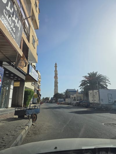 The image depicts a street scene in Hurghada, Egypt, viewed from a low vantage point, possibly from inside a vehicle. The sky is bright blue with a hint of sunshine. Buildings line the left side of the street, with storefront signage in Arabic. A tall minaret is visible in the distance, along the center of the road. Vehicles are parked on the right side of the street, adjacent to a palm tree. A small utility trailer is located on the edge of the sidewalk. The street itself is paved. The perspective is slightly skewed, making the scene appear tilted.