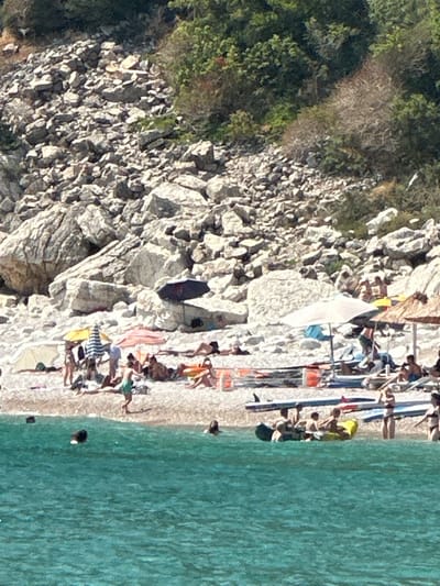 The image depicts a rocky coastline with a sandy beach next to turquoise water. The scene is populated by several people enjoying a sunny day. They are scattered along the beach, some lying on towels or sunbeds, while others are in the water. The time of day appears to be midday, judging by the strong sunlight and shadows.
Objects visible include beach towels, umbrellas, and what appear to be kayaks or surfboards lined up on the sand. The rocky terrain rises steeply from the beach, covered in sparse vegetation. The water is clear and inviting, with some small dark objects, possibly buoys or people swimming, visible in the distance.
The overall mood seems relaxed and summery, with people engaging in typical beach activities like sunbathing and swimming. The location is implied to be Mirište, Montenegro, a coastal area. There is no visible text in the image. The orientation of the image is rotated 90 degrees counter-clockwise.