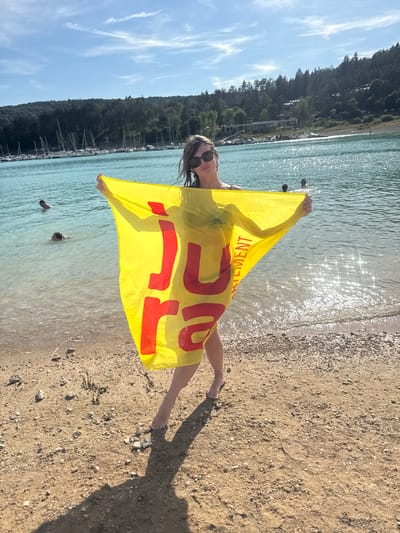 A woman stands on a sandy beach holding a yellow towel with red lettering that reads "JURA". The lake is calm and reflective, with several sailboats docked in the background. The woman is wearing sunglasses and a swimsuit. The sky is blue with scattered clouds. The location is the Lake Vouglans, near the city of Maisod, France.