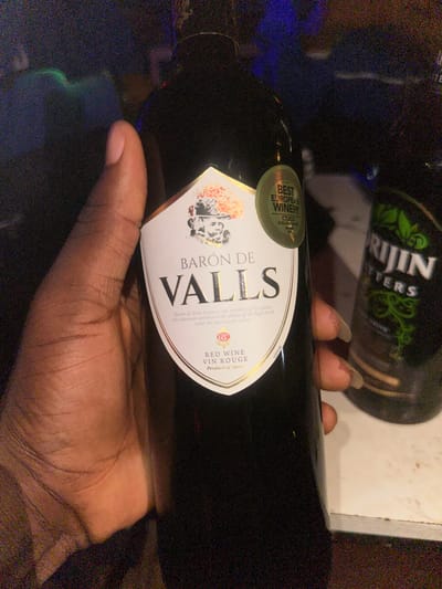 A close-up, eye-level shot shows a person's left hand holding a dark glass bottle of red wine. The wine bottle has a white label with the brand name "BARÓN DE VALLS" in large, dark lettering. Above the brand name are some decorative graphics and a circular sticker that reads "BEST EUROPEAN WINERY". Below the brand name, it states "RED WINE VIN ROUGE" and "Product of Spain". To the right of the wine bottle, a portion of another dark bottle with a green label is visible, which appears to be a beverage with the word "RIJIN" prominently displayed. The background is dark and out of focus, with hints of blue and green lights, suggesting an indoor setting at night. There is no indication of the city of Wumat, Nigeria, in the image.
