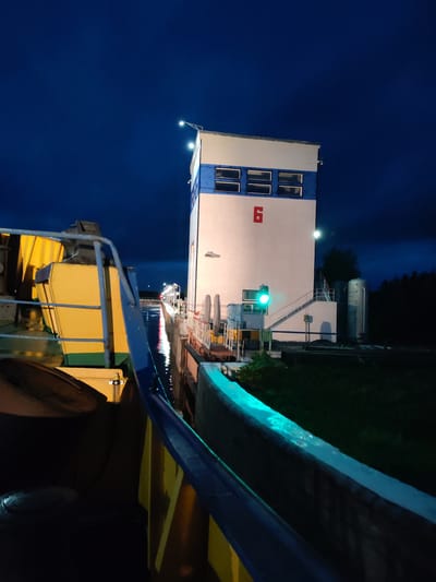 An exterior, low-angle, long shot captures a canal lock facility at night in Devyatiny, Russia. A large, yellow and blue barge occupies the foreground to the left. The barge's railing is visible, as is part of its deck. In the middle ground, a white and blue multi-story building, likely part of the lock control system, stands to the right. A green traffic light is illuminated on the side of this building. Stairs lead up the side of the building. A body of water, reflecting the lights from the building and distant lights, fills the space between the barge and the lock structure. The sky above is a deep, dark blue, indicating nighttime. The scene is illuminated by artificial lighting, casting reflections on the water and highlighting the architectural features of the lock. The background features a dark treeline.