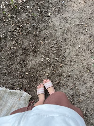 This media file shows a top-down view of a person's feet standing on the ground. The ground is composed of dirt, small twigs, and scattered leaves, suggesting an outdoor, natural setting. The person is wearing light pink and white striped sandals. Their right leg is visible, clad in a dusty rose colored garment, possibly a skirt or pants. A white fabric, possibly a bag or tarp, is partially visible in the lower right corner, also resting on the ground. The overall lighting suggests it could be daytime. The ground texture is rough and uneven, with some faint imprints possibly from previous footsteps. The setting appears to be a rural or wooded area in Вяззеўскі сельскі Савет, Belarus. No text is visible in the image. The focus on the feet and ground suggests a simple, grounded moment.