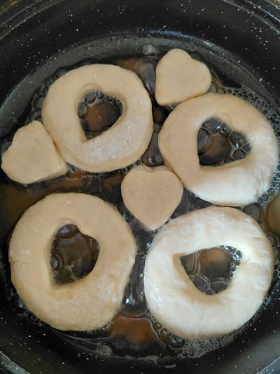 The image shows several raw donut shapes and heart-shaped pastries being fried in a black pan. The donuts have a light, doughy appearance and are in various stages of frying, with some showing bubbling oil around their edges. There are four ring-shaped donuts, each with a hole in the center, and two smaller heart-shaped pieces of dough. The oil is hot and actively bubbling, indicating the frying process is underway. The pan itself appears to be a non-stick surface, possibly a frying pan or a deep fryer. The lighting suggests it is daytime, and the close-up shot focuses solely on the food in the pan, offering no environmental context beyond the cooking vessel. No text is visible in the image. The scene depicts the preparation of a sweet treat, likely a dessert or snack.