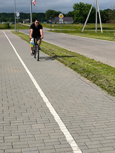 A man is cycling on a paved path next to a road. He is wearing a black t-shirt, black shorts, sunglasses, and sandals. He is carrying a multi-colored bag in the basket of his bicycle. The path is made of gray brick pavers with a white line marking the edge. The road is paved and has a grassy shoulder. In the background, there are trees, houses, and utility poles. The sky is cloudy, and the lighting suggests it is daytime. The overall scene appears to be in a rural or suburban area.