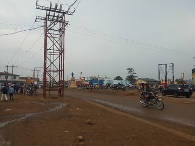 The image captures an unpaved urban intersection in Beni, Democratic Republic of the Congo, under an overcast sky. The foreground consists of reddish-brown dirt interspersed with muddy puddles and small rocks. A prominent, tall, red lattice-work power transmission tower stands left of center, supporting multiple overhead power lines that extend across the scene.

In the midground, a dirt road runs diagonally from left to right. A silver motorcycle with two occupants, both wearing dark clothing, travels rightward on this road. To its right, a dark-colored SUV is visible. On the left side of the road, approximately two dozen individuals are standing or walking, some in proximity to several parked motorcycles. Further into the scene, at what appears to be a central roundabout, a raised monument supports a blue-clad statue.

The background features various one and two-story buildings, some with visible storefronts, lining the perimeter of the intersection. Additional utility poles and power lines are present, crisscrossing the sky. On the far right, a large, bare metal framework, consistent with a billboard structure, stands against the horizon. The scene depicts active daily urban life.