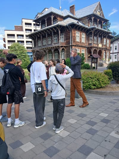The image depicts a group of people standing on a paved area in front of an old wooden building with intricate architectural details. A man in a suit and brown trousers is addressing the group, with his hand on his face as if adjusting his glasses. Several young people, appearing to be students, are listening attentively. The wooden building has a sign that appears to read "MUSEUM". In the background, a modern multi-story building is visible. The environment suggests an outdoor setting in a city, with trees and shrubs present. The overall scene is a guided tour or educational visit.