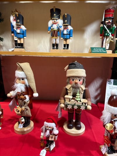 The image displays several wooden nutcrackers, likely for sale in a store. On the left, three traditional blue and black nutcrackers are displayed on a wooden shelf, alongside a sign noting, "To Purchase Please Ask An Associate for Help." To the right, on a red surface, are three nutcrackers with a folk art or woodland theme. One is in a brown hat, another in a tan cap with a burlap sack, and the third in a red Santa hat. Price tags, including numbers "101081-6" and "101151-1" and costs like "104.00," are attached to the figures. A sign with "Over 300 years ago wood turning" is also visible.