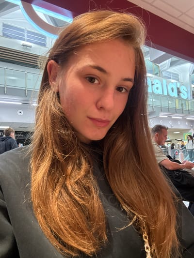 A young woman with long, reddish-brown hair is taking a selfie in an indoor environment, likely an airport or shopping mall, as indicated by the signage for "McDonald's" visible in the background. She is wearing a dark sweatshirt. Behind her, other people are visible, including a man seated at a table looking at a device and a woman standing near an escalator. The lighting suggests an overhead, artificial source. The location is not Pisa, Italy.