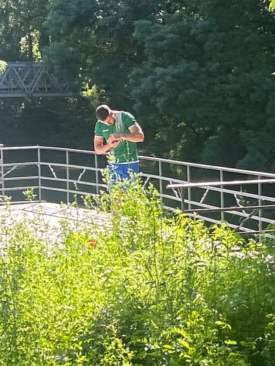 A man is standing in front of a metal structure, possibly a tower or support frame. He is wearing a green shirt with white accents and blue shorts. He appears to be looking down, possibly at a phone or other handheld device. The background includes dense green foliage and a partial view of a bridge or tower in the distance. The image was taken in Elhovo, Bulgaria.