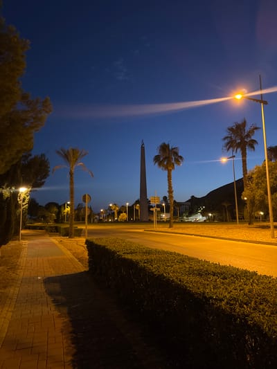The image depicts a street scene at dusk in Benidorm, Spain, featuring a prominent obelisk monument in the distance. The sky is a deep blue, transitioning to lighter hues near the horizon with a faint wispy cloud illuminated by distant lights. Tall palm trees are scattered throughout the scene, some silhouetted against the sky and others illuminated by streetlights. A well-lit pathway made of interlocking brick pavers runs along the left side of the image, bordered by manicured hedges. A street with a bright yellow asphalt surface is visible, with a few cars driving on it in the mid-distance. Streetlights cast a warm glow on the pathway and road, creating distinct shadows. The obelisk, a slender, pointed structure, stands tall in the background, appearing to be a significant landmark. The overall impression is a quiet urban environment illuminated by artificial light as night approaches.