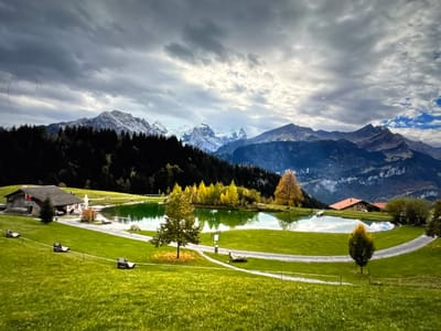 The image depicts a scenic mountain landscape with a pond in the foreground. The sky is overcast with dramatic clouds, hinting at a change in weather. In the mid-ground, a dark green forest lines the base of snow-capped mountains, which rise majestically in the background. The mountains display rugged terrain and snow-covered peaks.

In the foreground, a vibrant green meadow slopes gently downwards, dotted with several benches and some wire fencing. A small, man-made pond with still, reflective water is situated in the mid-ground, surrounded by lush green grass. A pathway curves around the pond. On the left side of the pond, a rustic wooden building with a dark roof and red accents is visible. Further to the right, another building with a terracotta-tiled roof is nestled into the landscape.

The foliage around the pond includes deciduous trees with yellow and orange leaves, suggesting an autumn season. A few lone trees stand in the meadow. The overall impression is one of natural beauty and tranquility, potentially a resort or a rural dwelling in a mountainous region. The image does not contain any people or specific indications of activity. The location is not Porrentruy, Switzerland, as there are no geographical features or man-made structures in the image that are recognizable as being from that specific city.