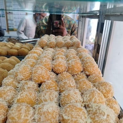 The image shows a display case filled with a large number of sweet pastries coated in shredded coconut. The pastries are stacked in rows in a glass display case. Two individuals are visible in the reflection, one wearing a white shirt and the other wearing a green and black shirt. The individual in the green shirt is holding a cell phone to take a photograph of the pastries. The location is a shop in Bhola, Bangladesh, at coordinates 22.686 latitude and 90.645 longitude. The pastries appear to be a popular local delicacy. The background is blurry, but it seems to be a commercial space.