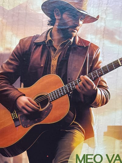 A man wearing a brown leather jacket and a cowboy hat plays an acoustic guitar. Part of the name "MEO VA" is visible in green on the right edge of the image. The location is likely Mèo Vạc Commune, Vietnam, based on the text fragment.