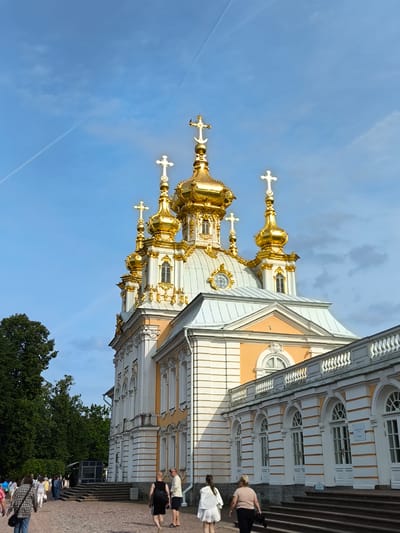 In Peterhof, Russia, on a bright, partly cloudy day, a grand Baroque-style building with multiple golden domes and crosses is prominently featured. The building's facade is a mix of white and yellow stucco, adorned with decorative elements and arched windows. A long colonnade with arched openings runs along the lower portion of the building. In the foreground, a cobblestone path leads toward the building, with a group of diverse individuals walking on it. Some are dressed casually, while others appear to be tourists. Trees and greenery are visible on the left side of the image, contrasting with the architectural splendor. The sky above is a clear blue with wispy white clouds and a faint contrail.