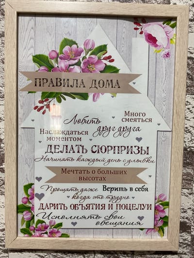 A framed, vertically oriented motivational poster is displayed against a grey, wood-paneled background. The poster features a house-shaped design with various handwritten and printed Russian text, adorned with floral illustrations of pink and purple flowers with green leaves.
The text includes:
- "ПРАВИЛА ДОМА" (House Rules) prominently at the top, on a banner.
- "Любить" (To love) and "Много смеяться" (To laugh a lot) in the upper section.
- "Наслаждаться каждым моментом" (Enjoy every moment) below.
- "ДЕЛАТЬ СЮРПРИЗЫ" (Make surprises) in larger font.
- "Начинать каждый день с улыбки" (Start every day with a smile) below that.
- "Мечтать о больших высотах" (Dream of great heights) on another banner.
- "Прощать, даже когда это трудно" (Forgive, even when it's difficult) followed by "Верить в себя" (Believe in yourself).
- "ДАРИТЬ ОБЪЯТИЯ И ПОЦЕЛУИ" (Give hugs and kisses) and "Исполнять свои обещания" (Keep your promises) at the bottom.
The poster is framed with a light-colored wood frame. The background behind the poster appears to be a textured surface, possibly a wall or fabric. No people or specific locations within Бескөл ауылдық округі, Kazakhstan are identifiable in this image.