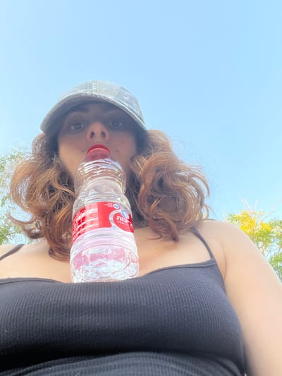 An adult female is depicted outdoors in the Brener Regional Council, Israel, in an upward-angle, eye-level shot. She wears a faded, patterned baseball cap and a dark, ribbed tank top with thin shoulder straps. Her reddish-brown, wavy hair frames her face. She is actively drinking from a partially filled clear plastic bottle of Mei Eden mineral water, identifiable by its red cap and red and white Hebrew label. The bottle is positioned at her mouth. The background consists primarily of a clear blue sky, with some green foliage visible at the extreme left and right, indicative of a bright, sunny outdoor setting.