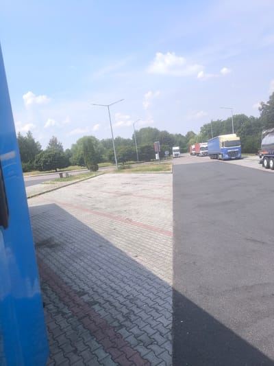 The image depicts a parking area for trucks under a bright, partly cloudy sky, suggesting it is daytime. The scene is an outdoor setting, likely a rest stop or service area given the presence of multiple parked trucks and paved surfaces. The foreground features a portion of a large blue truck, partially obscuring the view. Adjacent to the blue truck is a paved area made of interlocking concrete blocks, marked with red lines. This area is in shadow, indicating the sun's position.

Further into the image, a darker asphalt surface is visible, serving as the main road or parking lane for the trucks. Several semi-trailer trucks are parked in a line along this asphalt. The trucks vary in color and design, including a prominent blue and yellow DAF truck and a silver tanker truck. Green trees and foliage form a backdrop behind the trucks and the paved area, suggesting a location with natural surroundings. Tall lampposts are also present, typical of such roadside facilities.

In the distance, there are more trees and a hint of buildings or structures, but they are not clearly discernible. No people are visible in the image, and the overall impression is one of a quiet, functional location. The weather appears pleasant for travel, with a clear blue sky and scattered white clouds. The lighting and shadows suggest it is mid-day. The visible text on the DAF truck is "DAF" and a license plate which is illegible. The location context is Młyny, Poland.