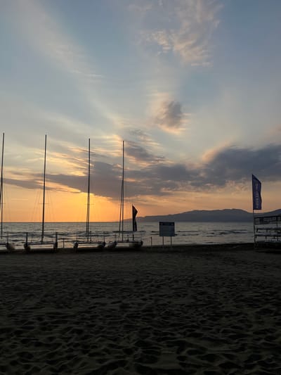 This image depicts a coastal scene at sunset, likely in the vicinity of Selçuk, Turkey. The foreground is dominated by dark, textured sand, showing subtle undulations and footprints. Stretching across the midground, several single-mast sailboats, appearing to be small catamarans, are lined up on the sand, facing the calm sea. Their light-colored hulls and dark mast silhouettes are visible against the water and the setting sun.
To the left of the sailboats, a white rectangular sign with dark text is partially visible, indicating a "SAILING CLUB." On the right side of the beach, a prominent blue vertical flag displays "AQUA SAILING" in white text. Another white sign, possibly related to "AQUA SAILING Lesson," is situated near the flag.
The background features the sea, which reflects the diminishing light, transitioning from dark near the shore to a lighter tone further out. A vibrant orange and yellow sunset illuminates the horizon line over the water, where the sun is obscured. To the right, a range of dark mountains is silhouetted against the colorful sky, extending into the distance. The sky above is a mix of pale blues, grays, and soft pinks, with scattered clouds, some appearing wispy and others denser over the mountains. No people or specific actions are discernible in the scene.