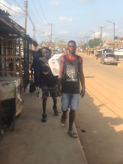 This image depicts a daytime street scene in Egan, Nigeria, under clear skies. Two male subjects are prominent in the foreground, walking on a concrete sidewalk. The subject on the right, facing the camera, wears a black sleeveless shirt with red trim, light blue denim shorts, and olive-green slide sandals. The subject behind and to his left, also facing the camera, carries a white cardboard box on his left shoulder. He wears a dark t-shirt, camouflage-patterned shorts, dark sturdy shoes, and a dark headwrap. Behind these subjects, another individual in a blue garment carries a dark bag.
To the left of the sidewalk are rudimentary market stalls constructed from wooden frames and corrugated metal, with some draped with fabric. The immediate right of the sidewalk features a wide, unpaved, light brown dirt road. In the mid-ground, a silver sedan or SUV is visible on the road, with multiple indistinct figures in the distance. The environment is characterized by numerous wooden utility poles supporting a dense network of overhead power lines. A communication tower is visible in the left mid-ground, beyond the market stalls. Various buildings with corrugated roofs line both sides of the street in the background. Shadows indicate direct sunlight.