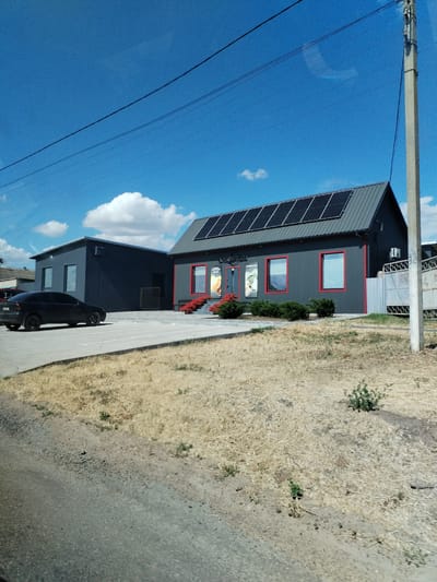 The image shows a modern, dark-colored building with a sloped roof covered in solar panels. The building has several windows with red trim. In front of the building, there are red flowers and green bushes. A black car is parked in the paved area in front of the building. To the left, there is another similar dark-colored building with two windows. The scene appears to be set in a somewhat rural or industrial area, with dry, yellowish grass in the foreground. Overhead, there are power lines and a clear blue sky with a few white clouds, suggesting a sunny day, likely midday. A concrete utility pole stands to the right of the main building. Some fencing is visible behind the building on the right. Text is visible on the front of the building, appearing to be a business name, but it is partially obscured. The overall impression is a contemporary commercial or industrial setting on a bright, clear day.