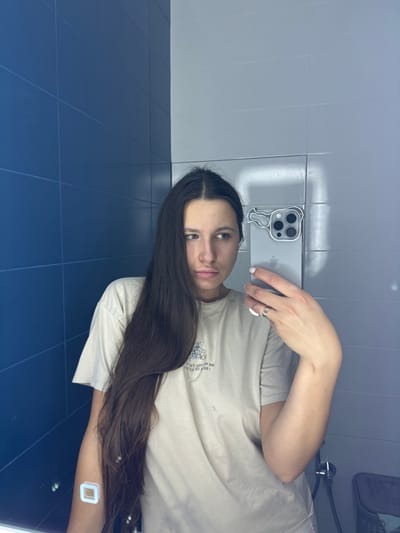 A female subject is captured in a mirror selfie within an indoor setting. She possesses long, dark hair, parted centrally, and wears a beige short-sleeved t-shirt featuring a graphic design on the chest. A square, white-outlined patch is visible on her left upper arm. Her right hand, adorned with a multi-banded silver ring on the ring finger and white-painted fingernails, holds a light-colored smartphone encased in clear plastic with a stylized animal head design around its triple camera module. The subject's expression is neutral, with her gaze directed to her left (viewer's left). The background consists of tiled walls: dark blue rectangular tiles are on the left side of the reflection, and light gray rectangular tiles are on the right, meeting at a vertical seam. A light source reflects off the lighter tiles. A portion of a dark, woven basket or container is visible in the lower right. The precise location within Shul'gino, Russia, is not ascertainable from the visual evidence.