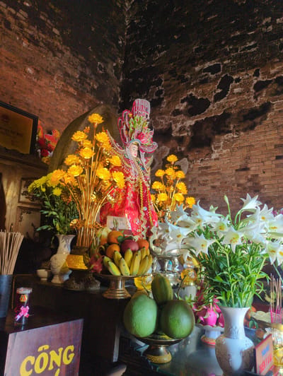 The image shows a shrine inside a structure with a brick wall. The shrine includes a statue dressed in red and adorned with yellow flowers, white lilies, and various fruits like bananas, oranges, and pomelos. There are also vases and a wooden sign with the word "CÔNG" written in yellow. The location is likely Tuy Hòa, Vietnam, given the Vietnamese text on the sign.