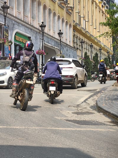 The image depicts a street scene in Sa Pa, Vietnam, under bright daylight. In the immediate foreground, two individuals are riding motorcycles away from the viewer. On the left, an individual operates a dirt-style motorcycle, heavily laden with gear bags attached to the rear. The rider wears a black and gray long-sleeved jacket, dark pants, and dirt bike boots, topped with a black helmet featuring red, white, and blue stripes. The motorcycle's rear license plate displays "97E-AK 037.50." To the right and slightly ahead, a second individual rides a dark-colored scooter or smaller motorcycle, wearing a blue jacket and a black helmet. Its visible rear license plate is "116.35."
The street is paved with asphalt, showing signs of wear, patches, and two circular manhole covers. A white lane line is partially visible in the mid-ground. To the far right, a tiled curb separates the road from a sidewalk.
On the left side of the street, a multi-story building features a gray, stone-textured lower facade. A prominent green sign for "TREKKING GEAR" is visible. Above this, the building transitions to a lighter color with a series of arched windows. Multiple ornate black streetlights, each with a hanging basket of pink flowers, are affixed to the building.
Further down the street, a white SUV is positioned in traffic, partially obscuring another scooter rider wearing a blue vest and a yellow helmet. A white Honda CRV is also partially visible on the left side, behind the lead motorcyclist. On the right, a long building with a distinctive yellow and cream facade extends into the distance, characterized by numerous arched windows and vertical architectural details. Parked motorcycles and pedestrians are visible along the right side of the street, including one individual in a light-colored shirt and hat. Green foliage from trees is visible on the far right. Shadows are cast across the street, consistent with sunny conditions.