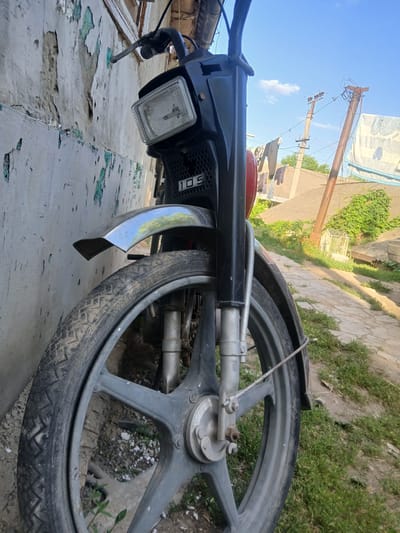 The image shows a close-up, angled view of the rear wheel and frame of a moped, leaning against a textured, weathered wall. The moped is black with some red accents visible on the body. The rear wheel is large with a grey, multi-spoke rim and a worn, black tire with a distinct tread pattern. Part of the exhaust pipe and a silver fender are also visible. The front of the moped, including a clear headlight and some black casing with textured perforations, is in the foreground, near the bottom left.

The background features a bright blue sky with a few wispy clouds. To the left and center, there are utility poles with wires, suggesting an outdoor, possibly rural or semi-urban setting. Green foliage and vegetation are visible in the mid-ground and behind the moped, alongside a paved or stone pathway. A white banner with some faint blue lettering is stretched across the top left, and laundry is hanging on a line between poles in the distance.

The overall impression is of a sunny day, as indicated by the clear sky and bright lighting. The weathered wall and the general appearance of the surroundings suggest a location that is not pristine, possibly a village or the outskirts of a town. There is no clear activity or event taking place; the moped appears to be parked.

A visible detail on the moped's casing, near the headlight, shows the text "105" in white. The image was taken in Pishchanka, Ukraine.