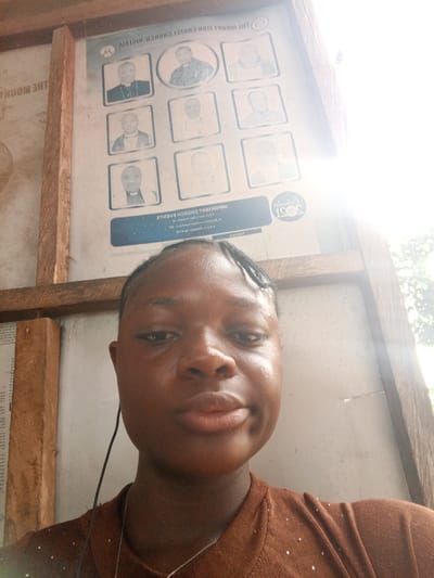 A young woman with dark skin and braids, wearing a brown shirt and headphones, stands in front of a display board attached to a wooden structure. The board features a title ("THE MOUNT ZION CHRIST CHURCH NIGERIA") and several portrait photographs of men. Below the portraits are text blocks. The surroundings suggest an outdoor location, possibly in Mbribit, Nigeria.