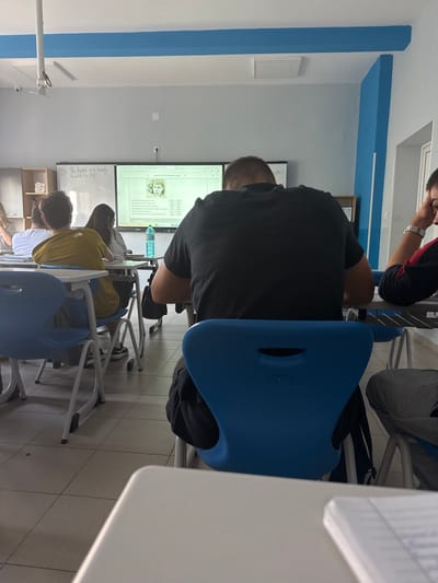 A classroom scene in Gorna Oryahovitsa, Bulgaria. Students are seated at desks, facing a whiteboard. Text and an image are displayed on the whiteboard. People are wearing casual clothing. Blue chairs are visible. The environment appears to be a typical classroom setup.