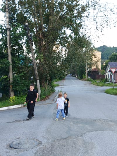 The image captures a street scene with three young boys. The boy on the left, wearing a black sweatshirt and pants, walks towards the viewer while looking to his right. He is holding a light green object in his right hand. Behind him, another boy, partially visible, is also walking in the same direction. To the right of the first boy, a second boy, wearing a white shirt with graphics and blue jeans, stands facing the viewer. Next to him, a third boy, dressed in a black t-shirt with a graphic and black pants, faces left. They are positioned on a paved road with some water stains and a manhole cover in the foreground. The road appears to be in a residential area, with trees and foliage lining the sides. In the background, there are houses, a tall building, and hills covered in trees. The sky is bright, suggesting daylight. The scene appears to be in Zlatoust, Russia, given the context provided.