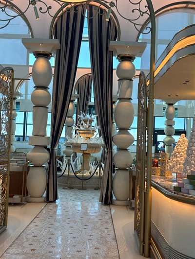 The image displays an interior scene, likely a hotel lobby or a high-end establishment, featuring ornate architectural details and decorative elements. The dominant colors are white, gold, and navy blue, creating a sense of opulence. Several large, white, urn-like structures with rounded, bulbous forms are positioned vertically, supporting golden railings and decorative scrollwork. These structures are interspersed with sections of navy and white striped fabric, possibly drapes or canopy elements.
In the background, a modern chandelier or art installation made of white, irregular shapes hangs from the ceiling, further enhancing the luxurious ambiance. There are glimpses of people in the background through glass partitions, suggesting it is an active public space. The floor is adorned with a patterned mosaic tile, adding another layer of intricate design. The lighting appears to be artificial, casting a warm glow on the surfaces. The overall impression is one of sophisticated interior design.
The scene appears to be set during the day, given the natural light filtering through what seem to be large windows or glass walls, though the primary illumination comes from interior lighting. No specific activity or event is clearly discernible, beyond the general presence of people and the implied use of the space as a reception or common area. The location context of Arnavutköy, Turkey, suggests a potentially waterfront or upscale urban setting. No visible text is present in the image.