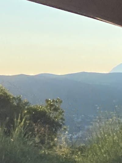 The image depicts a hazy, distant view of rolling hills or mountains at dusk or dawn. The sky is a pale yellow to white gradient, with a dark, blurred structure across the top edge of the frame, suggesting a foreground element such as a roof or balcony. The middle ground features layered, blue-toned mountain ridges that fade into the atmosphere. In the foreground, a dense cluster of green foliage, including bushes and tall grasses, partially obscures the view of the landscape and the settlement visible between the hills. The settlement appears as a collection of faint, indistinct shapes, suggesting houses or buildings. The overall impression is one of a natural landscape seen from an elevated vantage point, with a touch of human habitation interspersed. The provided location information for Seoce, Montenegro, indicates a coastal region with mountainous terrain, which is consistent with the visual elements presented in the photograph.