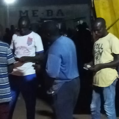 The scene depicts a dim-lighted evening gathering of four adult males in the city of Edopa, Nigeria. A building in the background displays partial white lettering "ME-BA", while a prominent yellow structure is visible on the far right. From left to right: a male in a striped shirt is partially visible. Adjacent to him, a male wearing a white t-shirt with a red and blue emblem and blue trousers looks downwards, appearing to handle small items in his hands. Centrally, a male in a plain blue t-shirt and dark trousers is observed from a rear angle. On the far right, a male in a light yellow t-shirt with "ROLL" printed text and light blue jeans is seen looking down, holding several small white rectangular objects, potentially paper or cards, in both hands. The individuals appear focused, engaged in an informal interaction, possibly involving a transaction or examination of items.