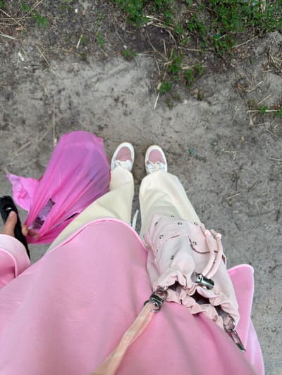 An individual is standing on a dirt path with sparse green vegetation and dried grass. They are wearing light-colored pants and a pink sweatshirt. On their left side, they are holding a black object, possibly a phone, and a bright pink plastic bag filled with items. On their right, a small, light pink drawstring bag with silver hardware is attached to their clothing. The person is also wearing white sneakers with pink accents. The scene appears to be outdoors in a casual, possibly rural or park-like setting.