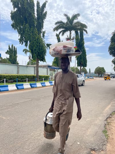 A man walks down a paved road in Unguwar Sarki, Nigeria, carrying a large metal container in his right hand and a tray of goods balanced on his head. The man is wearing a light brown, traditional outfit with a V-neck and short sleeves. His head is bowed slightly, and he is looking forward.

The tray on his head holds several items, including plastic-wrapped food items and a black container. The metal container in his hand appears to be a food carrier, with a silver lid and a cylindrical body with ventilation holes near the bottom.

The road is asphalt, with some cracks and unevenness. On the left side of the road, there is a white and blue striped concrete barrier separating the road from a grassy area with bushes and a light blue wall behind it. Trees, including palm trees and tall, slender trees with dense foliage, are visible in the background.

Further down the road, a silver car is parked on the left, and a yellow auto-rickshaw is moving away from the camera on the right. The sky is cloudy with patches of blue. The overall scene depicts a street vendor in an urban setting in Nigeria.