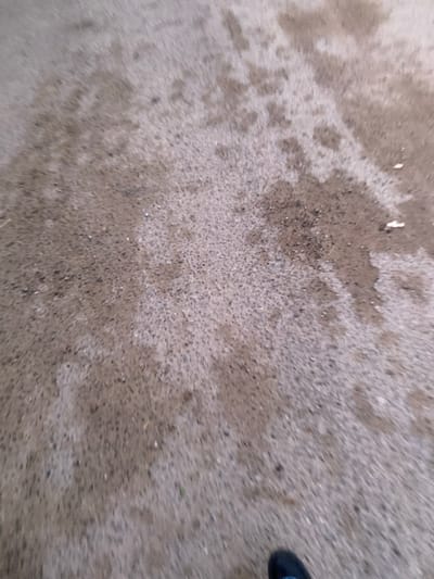 The scene, located in الغناشة, Tunisia, depicts a close-up, downward view of an outdoor ground surface. The ground consists of a granular, mottled material consistent with worn asphalt or rough concrete, presenting shades of light grey, brown, and dark grey. Irregular, darker patches suggest the presence of moisture or staining. Small, dark aggregate particles and sparse, lighter debris are scattered across the surface. In the lower right quadrant of the frame, the toe of a dark-colored shoe, likely black or dark blue, with a light-colored sole edge, is partially visible, indicating the presence of an individual. No other subjects or specific actions are discernible within the frame.