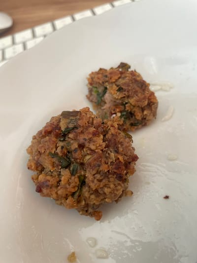 Two small, irregularly shaped, browned patties are visible on a white plate. The patties appear to be composed of ground meat and green herbs. The plate is slightly wet in some areas, with a few small droplets of liquid. In the upper background, a portion of a keyboard is visible, suggesting the scene is indoors. There is no indication of the city of Başakşehir, Turkey, within the image.