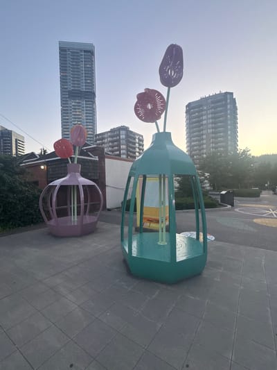 The image depicts two large, sculptural vases with stylized flowers, located in a public outdoor space in Coquitlam, Canada. The vase on the left is pink and appears to have a crown-like opening at the top, with green stems supporting three red flowers. The vase on the right is teal and has a similar opening, with green stems supporting two purple and one red flower. The sculptures are situated on a grey tiled walkway. In the background, several modern high-rise buildings are visible, indicating an urban setting. The sky is a pale blue, suggesting either early morning or late afternoon. The ground features colorful circular patterns. There are no people visible in the scene.