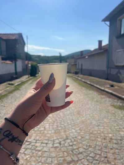 An outdoor, eye-level, medium shot shows a person's hand holding a white paper cup in the foreground. The hand has painted nails, one with a speckled pattern and another with red polish. A black string bracelet is visible on the wrist, along with a tattoo that appears to be script. The background depicts a cobblestone street lined with buildings on either side and a hilly, green landscape under a clear blue sky. The scene is set in Zelenikovo, Bulgaria, specifically on the street that leads towards the hills. The lighting suggests it is daytime, with the sun casting shadows and illuminating the sky.