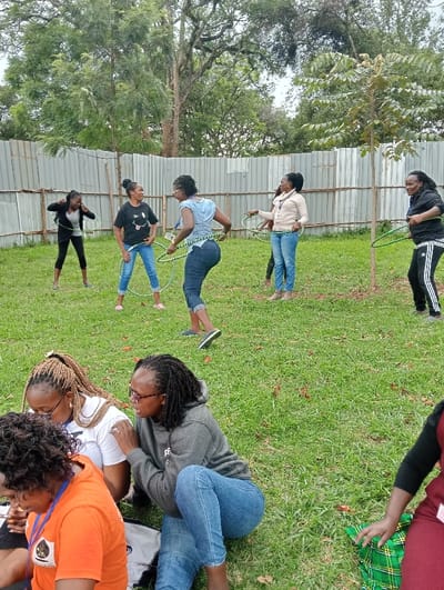 This outdoor image, set in Riara Ridge, Kenya, captures a group of women engaged in recreational activities on a grassy area. The scene appears to be daytime with overcast weather, suggested by the soft, diffused lighting and lack of strong shadows.
In the mid-ground and background, five women are actively hula-hooping. Most of them are smiling and appear to be enjoying the activity. They are dressed casually in jeans, t-shirts, and light jackets. One woman in a black t-shirt with a white smiley face emoji is laughing while attempting to hula-hoop. Another in a light blue top and jeans, wearing glasses, is also hula-hooping with a focused expression. To her right, a woman in a beige top and jeans holds a hula hoop, while further right, a woman in a black tracksuit and white-striped pants is also hula-hooping. A corrugated metal fence forms the immediate background, behind which numerous trees with dense foliage are visible.
In the foreground, three women are seated on the grass. One woman with braided hair, wearing a white t-shirt and glasses, is partially visible. Next to her, another woman with braided hair and glasses, wearing a grey hoodie and jeans (with "EF" or similar text partially visible on the hoodie), is looking towards her, possibly in conversation. On the far left, a woman in an orange t-shirt with a dark logo is also seated, facing away from the camera. A green and plaid fabric or blanket is visible on the grass next to the seated women on the right. The overall atmosphere appears relaxed and jovial, suggesting a casual gathering or team-building event.