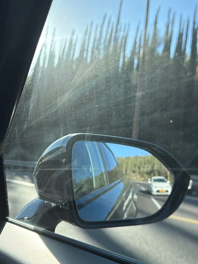 The image shows a view through a car window, looking at a side-view mirror. The mirror reflects a road with a white car driving ahead. Trees and a blue sky are visible in the background. The car window appears to have some dust or smudges on it, and the side of the car is visible in the foreground. The location is not discernible from the image itself, but the prompt specifies it is in the Mate Yehuda Regional Council, Israel.