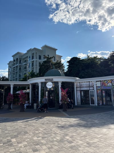 The image depicts a bright, sunny day in Alushta, Ukraine. In the foreground, a man is pushing a stroller across a paved courtyard. Behind him, a domed building houses a cafe named "POTOPAA" and a shop. The cafe features outdoor seating where several people are relaxing. To the right of the cafe are other shops, one with a sign in Cyrillic that appears to read "Unleagoe" and another with floral decorations. A modern, light-colored apartment building with multiple balconies is visible behind the shops, surrounded by lush green trees. The sky is a clear blue with scattered white clouds, and the sun casts shadows on the courtyard.
