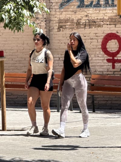 Two women are walking side-by-side on a sunlit pedestrian area in front of a brick wall in l'Hospitalet de Llobregat, Spain. The woman on the left is wearing a light-colored crop top with the number "92" on it, black shorts, and light-colored sneakers with pink socks. She has dark hair pulled back and is wearing sunglasses. She has tattoos on her arms and shoulders. The woman on the right has long dark hair and is wearing a black t-shirt, patterned leggings, and white sneakers with white socks. She is looking down and appears to be touching her face. Behind the women, there are wooden benches against the brick wall. A red stencil of the Venus symbol is visible on the wall to the right. The ground is paved with light-colored stones. There is a shadow from a leafy tree on the left side of the image.