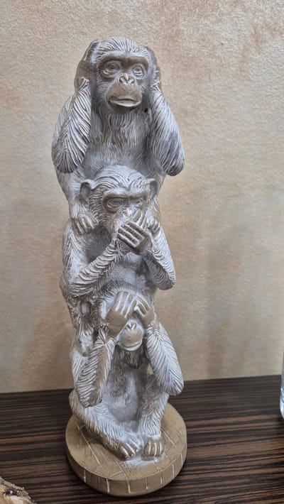 A stacked sculpture of three monkeys, each covering a different sense, is positioned on a wooden surface against a textured beige wall. The top monkey covers its ears, the middle monkey covers its mouth, and the bottom monkey covers its eyes. The sculpture is made of a light-colored material with darker accents to highlight details. The wooden surface has a dark, striped grain. The background wall has a rough, speckled texture. No people are present in the image. The scene does not provide any information about the city of Ravda, Bulgaria.
