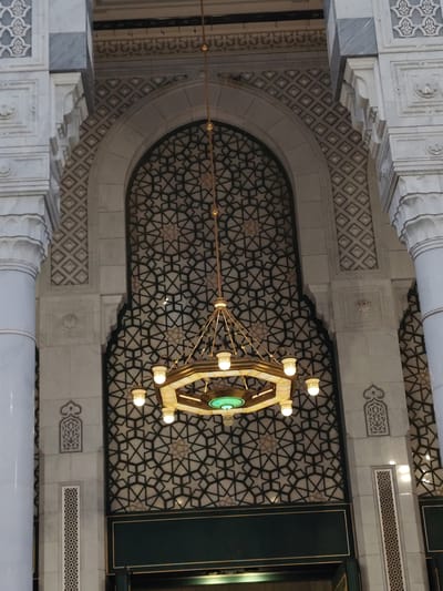 The image displays a grand interior architectural scene within a building in Makkah Al Mukarramah, Saudi Arabia, characteristic of Islamic mosque design. The central element is a large, ornate, gold-colored chandelier, likely made of brass or bronze, hanging directly in front of an expansive arched window or screen. The chandelier is octagonal, featuring multiple glowing bulbs around its perimeter and a distinct green-lit section at its base.
Behind the chandelier, the arched opening is filled with an intricate geometric pattern, consisting of interlocking star shapes and polygons in dark lines against a lighter background, creating a visually complex and elegant design. This geometric motif extends to the upper walls surrounding the arch.
The central arch is flanked by substantial, intricately carved columns and pillars made of light-colored marble or stone. These architectural supports feature various decorative elements, including textured patterns of small diamonds and squares, as well as stylized floral or arabesque carvings. The marble surfaces reflect the ambient light, enhancing the detailed craftsmanship.
Towards the bottom of the frame, a dark green panel, possibly part of a wall or a doorway, provides a strong color contrast to the light marble and golden chandelier. The overall aesthetic conveys a sense of grandeur, precision, and spiritual reverence through its elaborate decorative elements and majestic scale. No people or specific activities are depicted. There is no visible text.