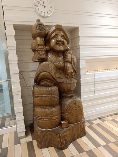 A large, light-brown, carved wooden statue of Daikokuten, a Japanese deity, occupies the central foreground. The smiling, rotund figure is depicted seated or leaning on two stylized wooden barrels or bales, holding a large mallet in its raised right hand and a bag in its left. The statue rests on an irregularly shaped dark brown wooden base. A small, light-colored wooden box is positioned on this base to the statue's right. The background consists of a light-colored wall featuring horizontal, recessed paneling, forming a wide alcove where the statue is placed. Above the statue, a round, silver-rimmed Seiko wall clock with a white face and black numerals displays approximately 9:55. To the left, a partial view of a vertical stack of white rectangular elements abuts a glass partition or door. The floor is tiled with alternating light and dark brown rectangular tiles arranged in a diagonal pattern. The environment is well-lit by artificial lighting. The specific location within Suginami, Japan, is not identifiable from the visual evidence presented in the image.