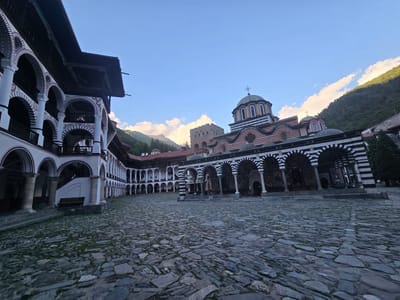The image depicts the central courtyard of the Rila Monastery, located in Rilski manastir, Bulgaria. The courtyard is paved with large, irregular stone slabs, some of which are covered in moss or grass. On the left side of the courtyard, a multi-story building with white walls and arched colonnades is visible. These arches are decorated with alternating red and white stripes. The building has a dark wooden roof. On the right side of the courtyard, a church with a prominent green dome topped with a cross is the focal point. This church also features arched colonnades with black and white striped arches and is adorned with colorful frescoes. Behind the church, a stone tower with a crenelated top is visible. In the background, a forested mountain slope rises under a clear blue sky with scattered white clouds. The sun is casting light on the upper parts of the mountains and the clouds. There are no people or animals visible in the image.
